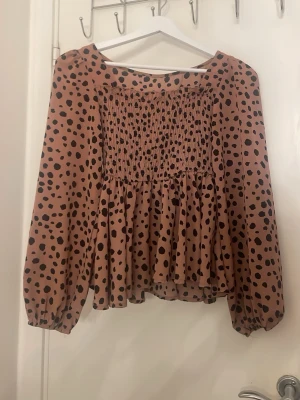 Brunprickig blus med smock - Söt blus i ljusbrun färg med svarta prickar och smockad överdel. Blusen har långärmade puffärmar med resår vid handlederna och en lös, flowy passform. Perfekt för en trendig och avslappnad look.