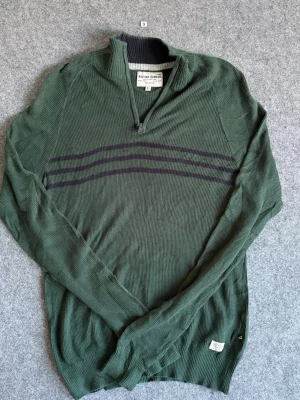 Grön tröja med dragkedja Nautica Jeans - Grön långärmad tröja från Nautica Jeans Co. med halv dragkedja och svarta ränder över bröstet. Tröjan har ribbad struktur och en liten etikett nertill. Perfekt för en avslappnad och stilren look.