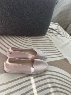 Ljusrosa ballerinaskor från Esprit - Säljer ett par ljusrosa ballerinaskor från Esprit med vit sula och diskret logga längs kanten. Skorna är i textilmaterial och har en enkel, klassisk design med rund tå och små detaljer i guld vid hälen. Perfekta för en stilren look.