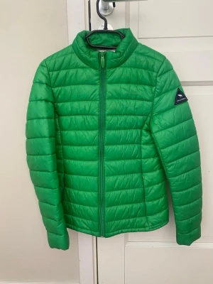 Grön pufferjacka från Lands' End - Snygg grön pufferjacka från Lands' End med quiltad design och dragkedja framtill. Jackan har en hög krage och en diskret logotyp på ärmen. Perfekt för vår och höst  och enkel att matcha med olika outfits.