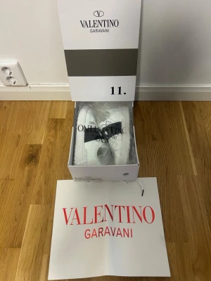 Valentino Garavani vita sneakers - Snygga vita sneakers från Valentino Garavani med svart band och nitar på sidan. Skorna har en chunky sula med mönster och är tillverkade i läder. Perfekt för dig som vill ha en exklusiv och trendig look.