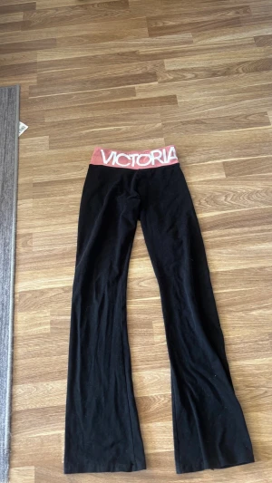 Svarta leggings från Victoria Sport - Snygga svarta leggings med utsvängda ben från Victoria Sport. Bred midjeresår i rosa med stor vit logga framtill. Mjukt och stretchigt material som sitter skönt på kroppen. Perfekta för en sportig och avslappnad stil. Storlek xs