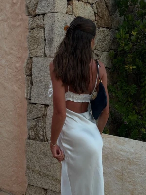 Vit långklänning i satin med snörning - Elegant vit långklänning i satin med smala axelband, djup v-ringning och snörning framtill. Klänningen har öppen rygg och en figurnära passform som ger en stilren och trendig look. Perfekt för sommarens alla tillfällen.