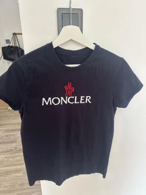 Svart tröja med Moncler logga - Snygg svart t-shirt med Moncler med vit broderad logotyp och röd detalj på bröstet. Klassisk rund halsringning och korta ärmar. Perfekt för en stilren och trendig look. 