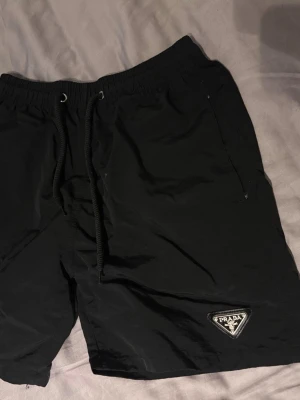 Svarta shorts från Prada med snörning - Svarta shorts från Prada med elastisk midja och snörning. Klassisk design med fickor och ikonisk Prada-logga på vänster ben. Perfekta för en stilren och avslappnad look.
