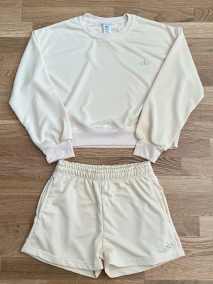 Beige loungewear set från Alo - Mysigt loungewear set från Alo i mjukt beige tyg. Setet består av en långärmad sweatshirt med ribbade muddar och matchande shorts med resår i midjan och fickor. Perfekt för chill dagar hemma. Diskret Alo-logga broderad på både tröja och shorts.