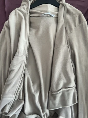 Beige hoodie med dragkedja lager 157 - Beige hoodie från 157 i mjukt fleecematerial. Modellen har dragkedja framtill, huva och två fickor på sidorna. Perfekt för chill dagar eller när du vill ha något bekvämt och stilrent. Enkel och clean design som passar till det mesta.