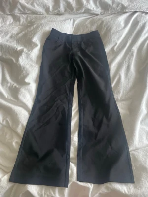 Svarta kostymbyxor med utsvängda ben - Snygga svarta kostymbyxor med utsvängda ben och klassisk passform. Byxorna har fickor bak och är tillverkade i ett slätt material som ger en stilren look. Perfekta för dig som vill ha en trendig och elegant siluett.