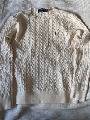 Vit kabelstickad tröja Polo Ralph Lauren - Kabelstickad tröja i offwhite från Polo Ralph Lauren med rund halsringning och ribbade muddar. Klassisk broderad logga på bröstet i mörkblått. Perfekt för dig som gillar tidlös och clean stil.