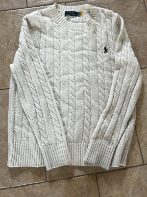 Vit kabelstickad tröja Polo Ralph Lauren - Påse ingår. Kabelstickad tröja i offwhite från Polo Ralph Lauren. Klassisk rund halsringning, ribbade muddar och nederkant samt den ikoniska broderade loggan på bröstet. Tröjan har en tidlös design och är gjord i mjukt material som känns skönt mot huden.