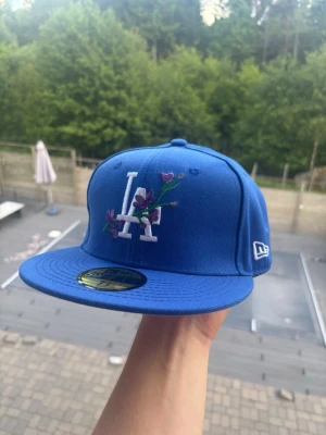 Blå LA-keps med broderade blommor - Snygg blå keps från New Era med LA-logga framtill och bak, dekorerad med broderade lila blommor på framsidan. Klassisk flat peak och broderad New Era-logga på sidan. Perfekt accessoar för att lyfta din streetwear-look.