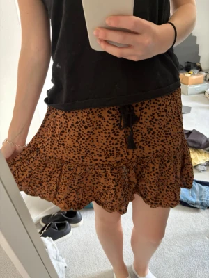 Brun leopardmönstrad kortkjol - Säljer en brun kortkjol med leopardmönster och volangkant. Kjolen har en svart snörning i midjan med tofsar som detalj. Perfekt för dig som vill ha en trendig och bekväm kjol med lite extra attityd.