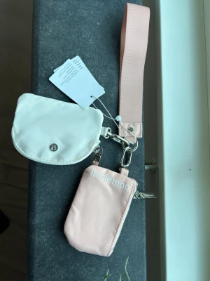 Lululemon plånbok och nyckelband - Säljer ett set med plånbok och nyckelband från Lululemon. Plånboken är ljusrosa med vit Lululemon-logga och den lilla fickan är vit med silverfärgad logga. Båda är tillverkade i ett slätt syntetmaterial och har metallspännen. Perfekt för kort och småsaker.