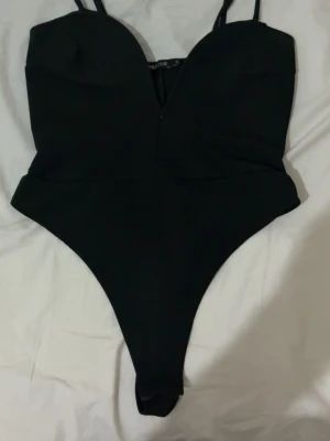 Svart body med djup urringning - Säljer en svart body från NLYone med smala axelband och djup v-ringning framtill. Bodyn har en figurnära passform och hög skärning vid benen. Perfekt att styla till jeans eller kjol för en trendig look.