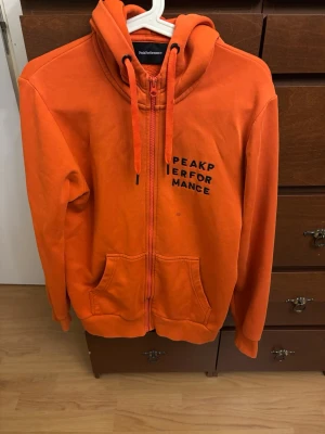 Orange hoodie från Peak Performance - Snygg orange hoodie från Peak Performance med dragkedja framtill och svart logga på bröstet. Tröjan har huva med dragsko och två stora fickor framtill. Tillverkad i mjukt bomullsmaterial, perfekt för chill dagar eller när du vill sticka ut.