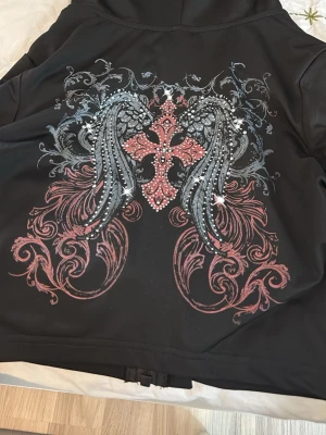 Svart hoodie med vingar och kors - Säljer en svart tränings hoodie med dragkedja och huva. På ryggen finns ett stort tryck med änglavingar, ett rosa kors och detaljer i grått och vitt. Hoodien har en cool och unik design som sticker ut. Perfekt för dig som gillar streetwear och detaljerade tryck.