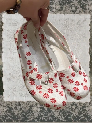 Vita ballerinaskor med röda blommor - Söta ballerinaskor i vitt tyg med röda blommor och ett dekorativt spänne över foten. Skorna har rundad tå och låg sula, perfekta för en lekfull och trendig look. Lätta att matcha till olika outfits och riktigt bekväma att bära hela dagen.