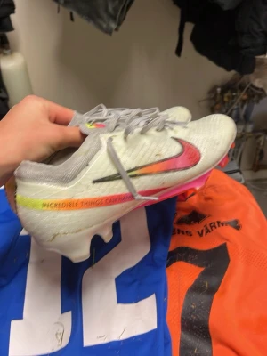 Nike vita Mercurial fotbollsskor - Säljer ett par vita Nike Mercurial fotbollsskor med färgglad swoosh i rosa, orange och svart. Skorna har texten 'INCREDIBLE THINGS CAN HAPPEN' på sidan och sulan är neonrosa med inslag av orange och grönt. Lätta och smidiga skor i syntetmaterial, perfekta för snabba spelare. 