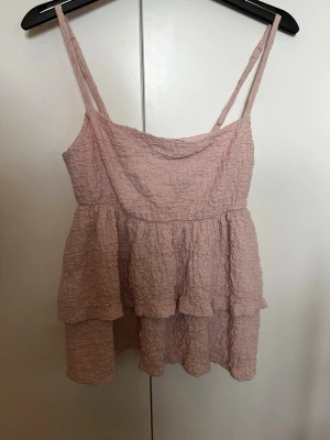 Ljusrosa volangtop från Gina Tricot - Supersöt ljusrosa topp från Gina Tricot med tunna axelband och volangdetaljer. Toppen har en crinklad struktur och är perfekt för varma dagar. Bra skick. Säljer då den inte kommer till användning.
