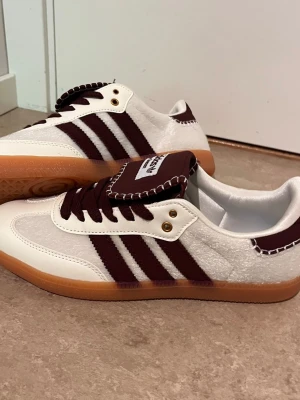 Adidas Samba OG vit och vinröd - Säljer ett par Adidas Samba OG sneakers i vitt och vinrött med klassiska tre ränder på sidan. Skorna har en låg profil, gummisula och detaljer i både mocka och läder. Tungan är extra bred med kontrastsöm och Adidas-logga.