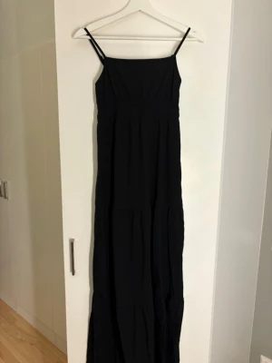 Svart långklänning med tunna band - Säljer en svart långklänning med tunna axelband och rak urringning. Klänningen har en enkel och stilren design som faller snyggt längs kroppen. Perfekt för dig som gillar minimalistisk stil och vill ha en klänning som funkar till många tillfällen.