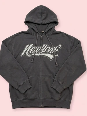 Mörkgrå hoodie med tryck och dragkedja - Säljer en mörkgrå hoodie med vitt tryck över bröstet. Det står NewYork. Tröjan har huva med snörning, dragkedja framtill och stora fickor. 