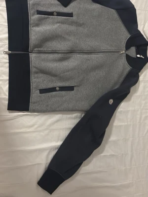 Grå och marinblå hoodie från Moncler - Snygg hoodie från Moncler i grått och marinblått med dragkedja framtill och två fickor med knappdetaljer. Ärmarna är marinblå och har Moncler-loggan på vänster ärm. Tillverkad i mjukt sweatshirtmaterial, perfekt för en avslappnad och trendig look. Det finns en sån knapp som e sönder man kan inte alls se de om man har på sig för för att de är tekt som man ser på bilden och de finns fläckar som man kan knappt se men jag tror att de går bort med tvätten och tröjan är äkta!!!