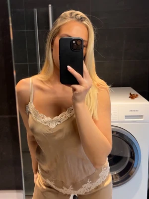 Pyjamasset från Intimissimi - Säljer ett set med beige satinpyjamas bestående av linne och shorts. Linnet har tunna axelband och vacker vit spets längs urringning och nederkant. Shortsen har matchande spets och dragsko i midjan. Perfekt för mysiga kvällar hemma 🩷