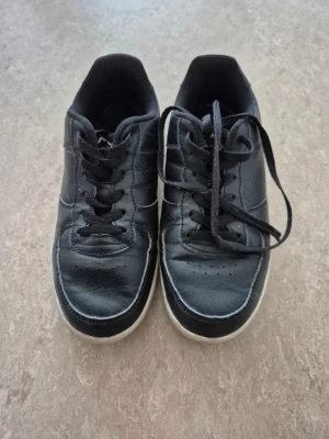 Sneakers - Klassiska svarta sneakers från Nonation med vita sulor och svarta skosnören. Skorna har en låg profil och är tillverkade i syntetmaterial med diskreta sömmar och perforeringar på tån för extra ventilation.