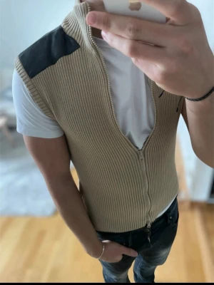 Beige väst - Säljer en beige stickad väst från smog med dragkedja framtill och svarta detaljer på axlarna. Västen har ribbad struktur och hög krage, perfekt att bära över en t-shirt för en cool och avslappnad stil. Mycket bra pris och väldigt snygg väst så passa på! Priset kan ändå diskuteras vid snabb affär.