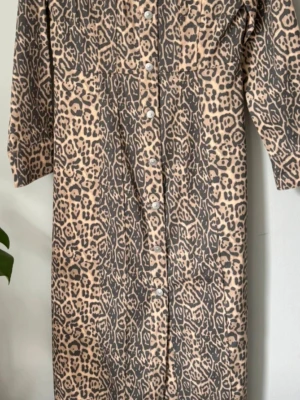 Leopardmönstrad långklänning med knappar - Leopardmönstrad långklänning i beige och svart med långa ärmar och knappar framtill. Klänningen har en avslappnad passform och är perfekt för dig som vill sticka ut. Materialet känns mjukt och följsamt.