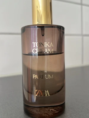 Zara Tonka Cream Eau de Parfum - Tonka Cream Eau de Parfum från Zara i en elegant cylinderformad glasflaska med guldigt lock. Flaskan har en mjuk beige-brun ton och text i guld. Doften är krämig och söt, perfekt för dig som gillar varma och mjuka parfymer.