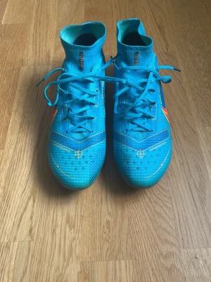 Blå Nike Mercurial fotbollsskor - Säljer ett par blå Nike Mercurial fotbollsskor med högt skaft och snörning. Skorna har orange och gula detaljer samt Nike-logga på sidan. Tillverkade i syntetmaterial för bra grepp och kontroll på planen. Perfekta för dig som vill sticka ut på fotbollsplanen. 