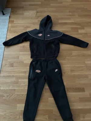 Svart Nike mjukisset med hoodie - Svart mjukisset från Nike med matchande hoodie och byxor. Hoodien har dragkedja, huva och rosa Nike-logga på bröstet. Byxorna har resår i midjan, sidofickor och rosa Nike-logga på låret. Perfekt för chill eller träning. Mjukt material som känns skönt mot huden.