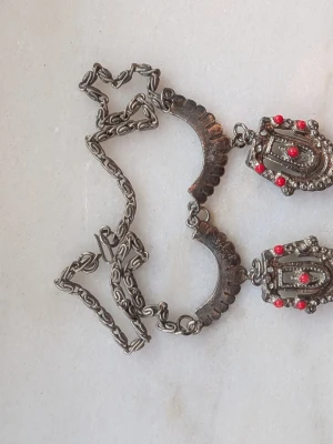 Vintage halsband med röda pärlor - Unikt halsband i antik silverfärg med detaljerade länkar och två stora hängsmycken dekorerade med små röda pärlor. Halsbandet har en bohemisk och rustik känsla med vackra mönster på metallhänget. Perfekt för dig som gillar statement-smycken.
