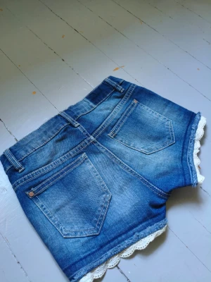 Blå jeansshorts med spetskant - Snygga blå jeansshorts med vit spetskant längst ner. Klassisk femficksmodell med knapp och dragkedja framtill. Shortsen har en tvättad look och är perfekta för varma dagar. Passar dig som gillar en söt men avslappnad stil. 