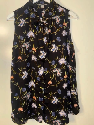 Svart blommig ärmlös blus med knyt - Snygg svart blus från H&M med färgglatt blommönster i blått, lila och orange. Blusen är ärmlös och har en dekorativ knytning vid halsen. Tillverkad i ett lätt och luftigt material som ger ett flowy intryck. Perfekt för dig som gillar en feminin och trendig stil.