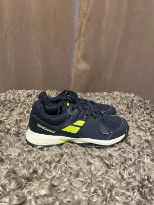 Mörkblå tennisskor från Babolat - Snygga mörkblå tennisskor från Babolat med gula detaljer och vit sula. Storlek eur 33.Skorna har klassisk snörning, perforerade partier för ventilation och en stabil design som passar perfekt för intensiva matcher på banan. Tillverkade i slitstarkt syntetmaterial. 