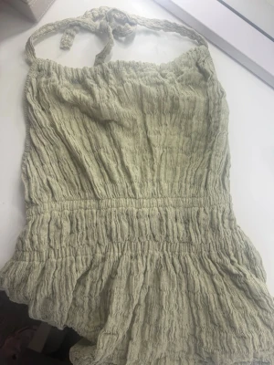 Grön tröja med knytband - Supersöt ljusgrön jumpsuit i crinklat tyg med smockdetaljer och knytband i ryggen. Axelbandslös design med volangkant nertill och öppen rygg. Perfekt för sommardagar och enkel att styla med sneakers eller sandaler.