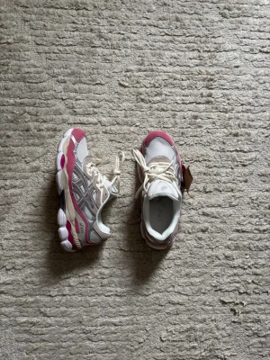 ASICS vita och rosa sneakers - Snygga sneakers från ASICS i vitt och rosa med grå detaljer. Skorna har meshpaneler för bra ventilation, klassiska snörningar och en chunky sula med GEL-dämpning. Perfekta för dig som gillar sportig stil och bekväma skor.