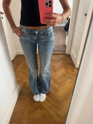 Ljusblå bootcut jeans med låg midja - Säljer ett par ljusblå bootcut jeans med låg midja och klassiska fem fickor. Jeansen har slitna detaljer och en avslappnad passform. Perfekta för en trendig och avslappnad look. Materialet är denim i bomull.