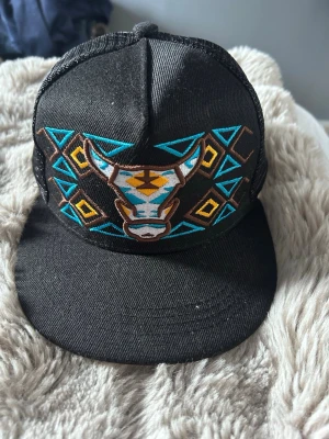 Svart keps med broderad tjur och mönster - Säljer en svart keps med platt skärm och mesh på sidorna. Framtill finns ett broderat motiv av ett tjurhuvud i turkos, brunt och vitt, omgiven av geometriska mönster i gult, blått och brunt. Perfekt för dig som gillar unika accessoarer.
