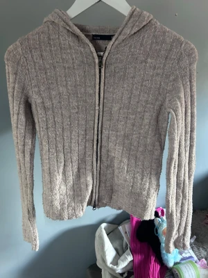 Beige ribbad hoodie med dragkedja - Mysig beige hoodie med ribbat mönster och dragkedja framtill. Tröjan har huva och är tillverkad i ett mjukt, stickat material som känns skönt mot huden. Perfekt för dig som gillar en avslappnad och stilren look.