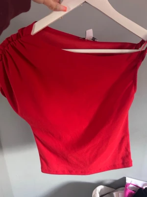Röd oneshoulder topp med rynk - Cool röd oneshoulder topp med rynkade detaljer på axeln. Toppens asymmetriska snitt ger en unik look och den är tillverkad i mjukt bomullsmaterial. Perfekt för dig som vill sticka ut och ha något trendigt i garderoben.
