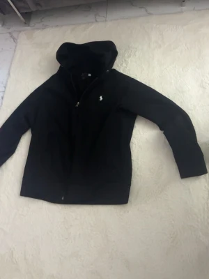 Svart hoodie från Polo Ralph Lauren - Klassisk svart hoodie från Polo Ralph Lauren med dragkedja framtill och vit broderad logga på bröstet. Hoodien har huva och långa ärmar, perfekt för en avslappnad och stilren look. Tillverkad i mjukt material för extra komfort.