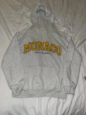 Grå hoodie med MONACO-tryck - Säljer en grå hoodie med gul MONACO-text och svart 'fitness & athletics' tryckt under. Klassisk känguruficka framtill, ribbade muddar och justerbar huva. Perfekt för chill dagar eller när du vill ha en sportig look. Älskar verkligen hoodien men tyvärr kommer den ej till användning 💕