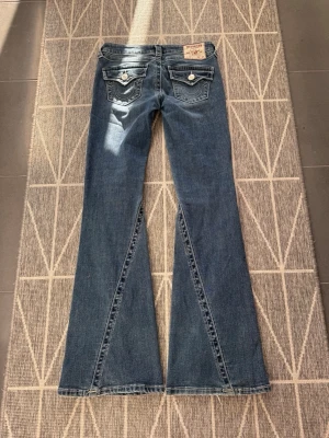 Blå bootcut jeans från True Religion joey bootcut low rise flare - Säljer ett par blå bootcut jeans från True Religion med slitna detaljer framtill och snygga fickor bak med dekorativa sömmar och knappar. Jeansen har låg midja och klassisk femficksmodell. Perfekta för dig som gillar en avslappnad men trendig stil. w25 bootcut low rise flared (xs)