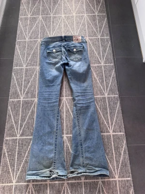 Blå bootcut jeans från True Religion joey low rise flare  - Säljer ett par ljusblå bootcut jeans från True Religion med slitna detaljer vid benslut och bakfickor med lock och knappar. Klassisk femficksmodell i denim med snygg tvätt och broderade detaljer på bakfickorna. w24 joey low rise flare bootcut flared 