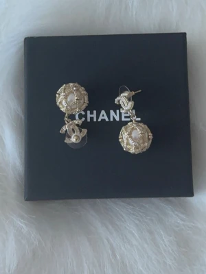 Chanel örhängen med pärldetaljer - Lyxiga Chanel örhängen i guldton med dekorativa pärlor och ikoniska CC-loggan. Örhängena har en rund form med intrikata detaljer och pärlor i mitten, vilket ger en elegant och exklusiv känsla. Perfekta för dig som vill sticka ut med klassiska accessoarer.