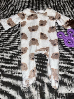 Vit pyjamas med nallemönster - Supermjuk vit pyjamas i fleece med gulligt brunt nallemönster över hela plagget. Dragkedja framtill för enkel påklädning och inbyggda fötter för extra mysfaktor. Perfekt för att chilla hemma eller sova bekvämt.